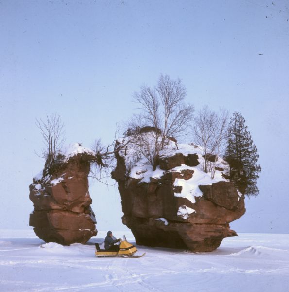 View across frozen bay towards a person riding a snowmobile in front of a split rock formation on Chequamegon Bay.