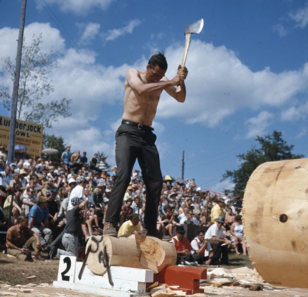 A shirtless man is standing on top of a log ready to chop a log with an axe. A crowd of people are watching from stands behind him.