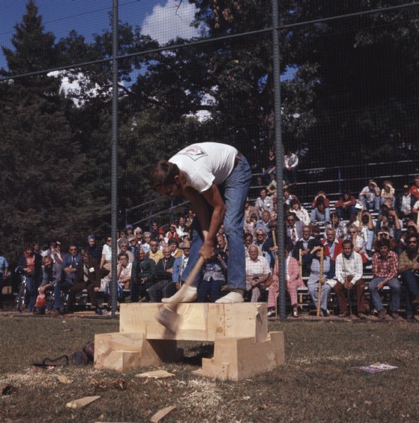 A man is standing on top of a braced wood block. He is bending over as he swings his ax at the wood block on which he is standing. A crowd of people are watching from the stands behind him.
