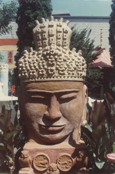"Monumental Oriental Head" View of a large, faded concrete-cast "Monumental Oriental Head" of the Buddha with heavily textured light-colored hair and topknot. With dimensions of 110" x 45" x 78," the piece is sitting on a base ornamented with Zodiac symbols (with Leo and Scorpio in front) in Sid Boyum's backyard. Sculpted owls are sitting beneath the bust on the base. The brick building of the Madison-Kipp Corporation is in the far background.