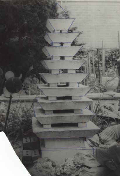 Unfinished cast-concrete sculpture identified as "Multi-tiered Pagoda" and measuring 92" x 32" x 32." It is standing in Sid Boyum's backyard. The brick wall of the Madison-Kipp Corporation is in the far background. 
This black-and-white photograph shows blue ballpoint-pen ink lines outlining the edges of the tiered squares that decrease in size from the base to a missing pyramidal point.
A handwritten notation of Sid's on the back of an undigitized image states that the pagoda took two-and-a-half tons of cement to make. 
