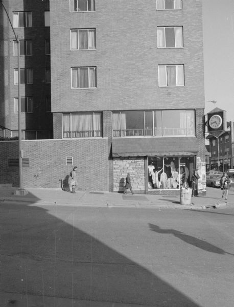 Manchester Corner Store and Student Apartments View across North Frances Street towards a student apartment building at the corner of State Street. On the ground floor is the Manchester Corner Store, with a show window displaying clothing. Posters cover the brick corner of the building next to the show window. A clock is hanging above the sidewalk at the corner.