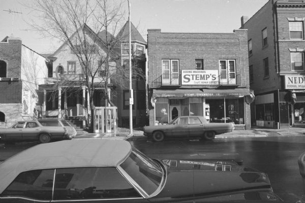 The Dollard Home and Harry Wigonitz Building View across State Street towards the Dollard Home and the Harry Wigonitz Building. The Dollard Home is a Queen Ann-style house with porch and turret. Two phone booths are on the sidewalk in front. The Harry Wigonitz Building contains Stemp's Typewriter Co., with typewriters on display in the show window. Cars are parked on both sides of the street. To the left of the house is Gino's, and to the right of Stemp's is Nedrebo's Formal Wear.