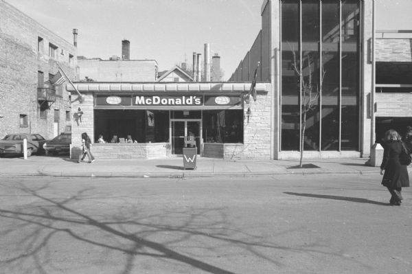 McDonald's View across North Lake Street towards a McDonald's fast food restaurant. A woman is walking across the street on the right, and two young women are walking by the McDonald's on the sidewalk as a man is opening the door to go inside. Two flags are hanging from the building, one of them an American flag.