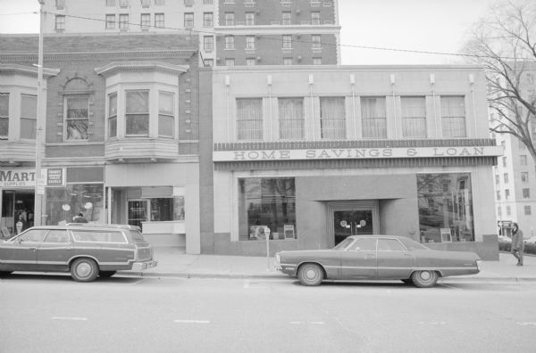 Home Savings & Loan Bank View across South Carroll Street, at the intersection with West Washington Avenue, towards the Home Savings & Loan Bank. To the left of the bank is part of the Badger Block Annex. Pedestrians are walking on the sidewalk. Two cars are parked along the curb. The windows of the Home Savings & Loan Bank are reflecting the Wisconsin State Capitol which is across the street.