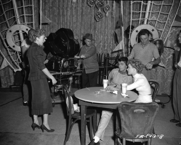 Ida Lupino, Keefe Brasselle and Sally Forest Director Ida Lupino is standing on the left, while actors Keefe Brasselle and Sally Forest are sitting at a table on the set of the 1949 film "Never Fear." A movie camera is being set up in the background.