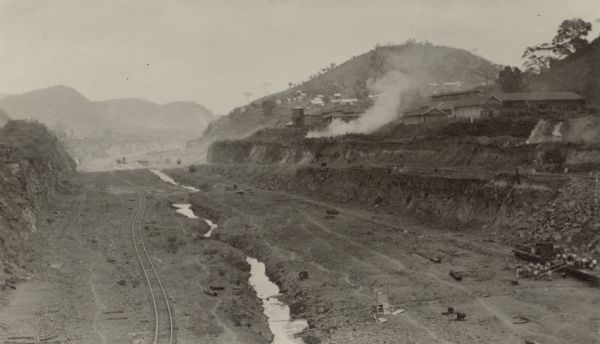 Culebra Cut An elevated view of the vast excavation of the Culebra Cut during construction of the Panama Canal. Railroad tracks and a stream are running through the cut. On the hillside above are large sheds and a water tower, with houses on the hill in the background.