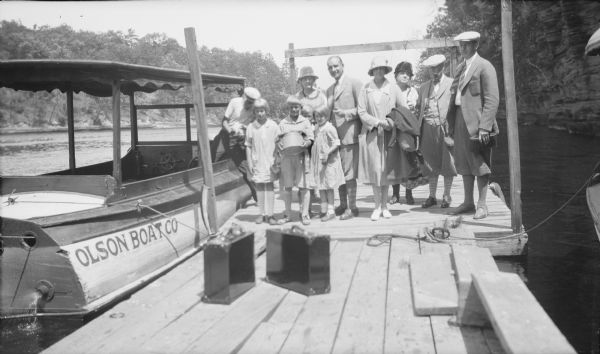 At the Dells Members of the extended Brumder family pose on a pier at the Wisconsin Dells. A motor launch with "Olson Boat Co" painted on the side is moored at the pier. Herbert E. Brumder holds a large round tin box as his brother Philip looks over his shoulder. At Herbert's right is his first cousin once removed Mary Messinger; her sister Joanne Messinger is at his left. Behind them, from left, are an unidentied man stepping into the boat; unidentied woman; Clifford Messinger holding a pipe; Margaret Bouer Brumder with a coat over her arm; the rest are unidentified. The men are wearing knickers with patterned socks. Two metal suitcases sit on the pier.