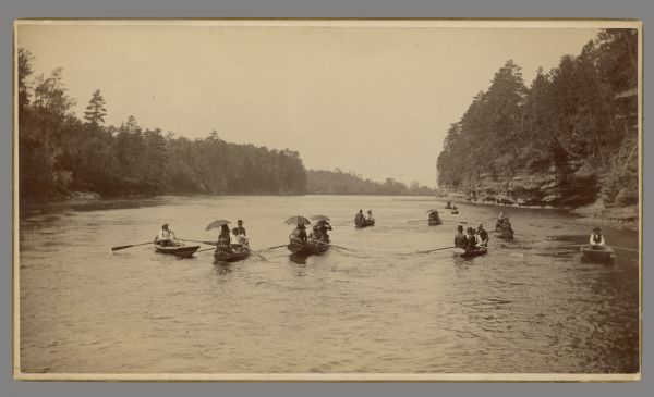 Fleet of rowboats carrying men and women with umbrellas.