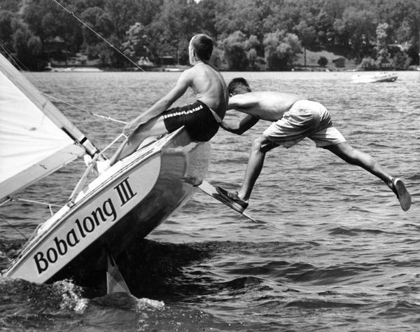 Two boys on the class-C scow sailboat "Bobalong III," with one boy balancing off the side while standing with one leg on the bilge keel.
