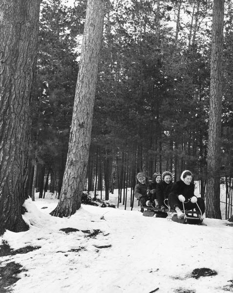 Three women and a man slide down a snowy hill on a wooden toboggan.
