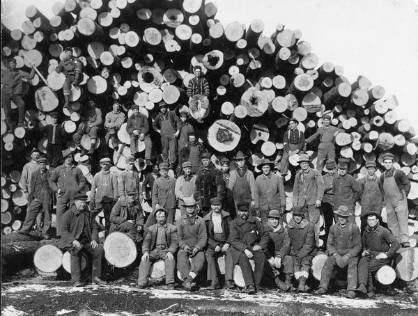 Thirty-nine lumberers posed in front of a large stack of cut logs.