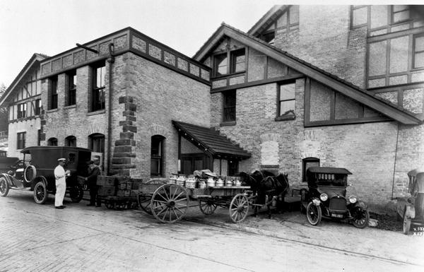 Exterior view of a dairy. Two cars and a horse-drawn wagon loaded with milk cans are parked in front of building.