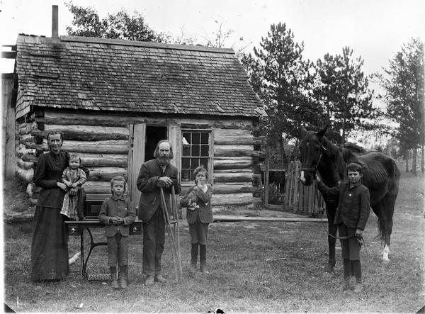Farm family posing in front of a log home. There is a man on crutches in the center, a woman is holding a small child standing on a table, and two children standing, one holding a cat. On the right is a boy is standing and holding the bridle of a horse.
