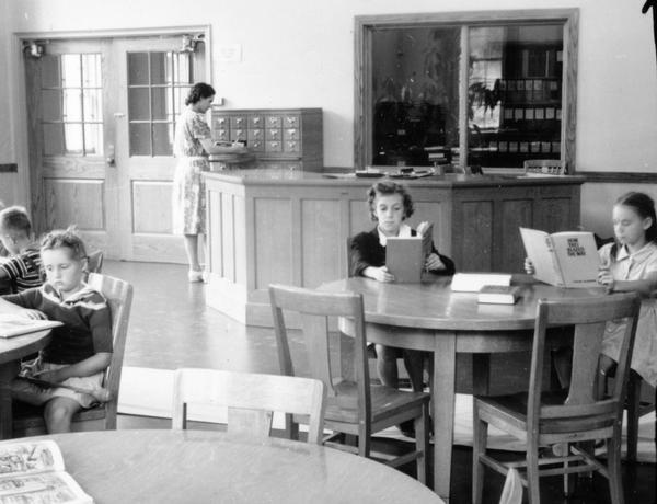 Young children reading at the library. A librarian works near the circulation desk in the background. When it opened in 1938, the Library had a seating capacity of 72 and a 5600 volume capacity, though it only had 2000 volumes at the time.