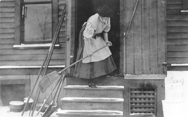 Woman sweeping snow off a porch. She is wearing a dress and apron.