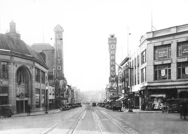 State Street view looking toward the University of Wisconsin, with the 200 block in the foreground. The marquee for the Capitol Theatre reads "'Jealousy,' All Talking," and a sign below the Orpheum Theatre's marquee reads: "Thanksgiving Month." Other storefronts include Rennebohm's Drugstore, Hills Dry Goods Co., and Kessenich's. In the far background is Bascom Hill of the University of Wisconsin.
