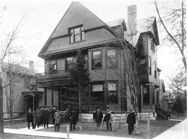 Men standing outside of the Delta Upsilon fraternity house at 673 State Street, at the intersection of State and Lake Streets. Lawrence Whittet stands to the left of the bicycle and Burt Shirley is on the far right.
