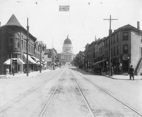 View looking up the 200 block of State Street from West Johnson Street, showing the Wisconsin State Capitol dome under construction. The bank on the left is a branch of the Bank of Madison. An overhead sign says the speed limit is 8 miles per hour.