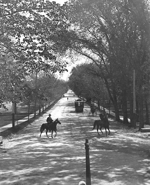 Tree-lined State Street from Bascom Hill on the University of Wisconsin-Madison campus. Traffic on State Street includes two well-dressed gentlemen on horseback, and a streetcar. The Wisconsin State Capitol is in the far distance.