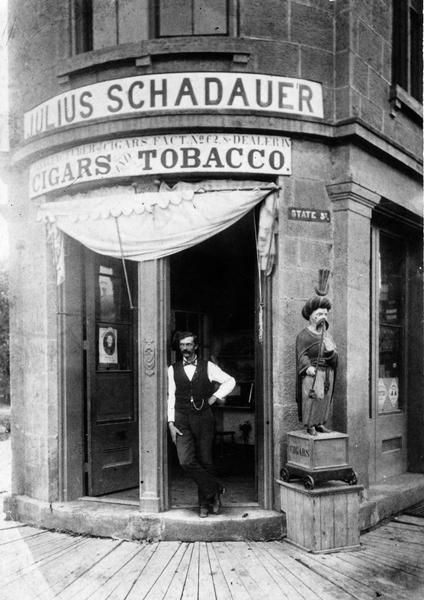 A man posing in the open doorway of Julius Schadauer's tobacco shop at 101 State Street. The sign above the doorway reads: "Schadauer of Cigars Fact. No 62, & Dealers in Cigars & Tobacco." To the right of the doorway is a cigar store statue depicting a man with a long moustache wearing a turban with a crescent symbol on his head and smoking a long pipe. The sculpture is standing on a box with wheels, and the sign painted on the box reads: "Cigars."