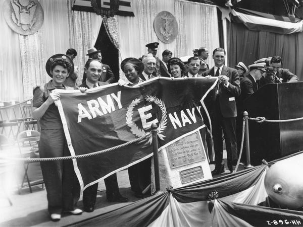 Group of McCormick Works employees holding up Army-Navy "E" banner with torpedo in foreground. The McCormick Works was built by Cyrus McCormick in 1873 and became part of International Harvester in 1902. The factory was located at Blue Island and Western Avenues in the Chicago subdivision called "Canalport." It was closed in 1961.