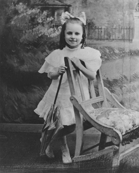 Agnes Moorehead as a child, posing with a parasol next to a chair in front of a photographic studio painted backdrop. She is wearing a white dress and has a bow in her hair.