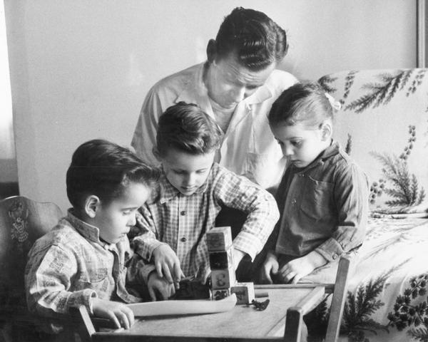 A father, Peter Froeming, watching as his three children George, Robert, and Roseann are playing with wooden blocks, with a small toy canoe in the foreground.