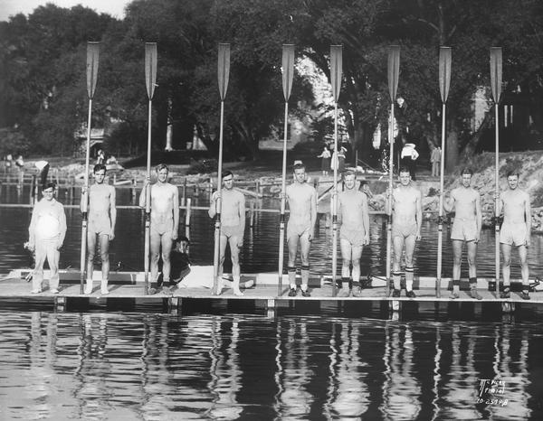 University of Wisconsin-Madison crew team holding their long oars, with coxswain on a pier on Lake Mendota.