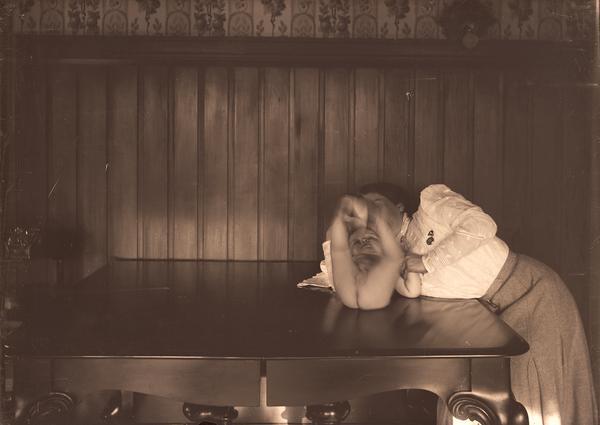 A baby lies on the dining table playing with his feet while his mother looks on.

