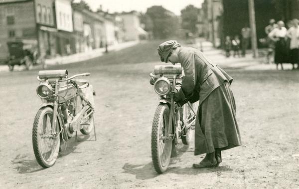 A woman adjusts her motorcycle in the middle of a street. A motorcycle is parked next to her.