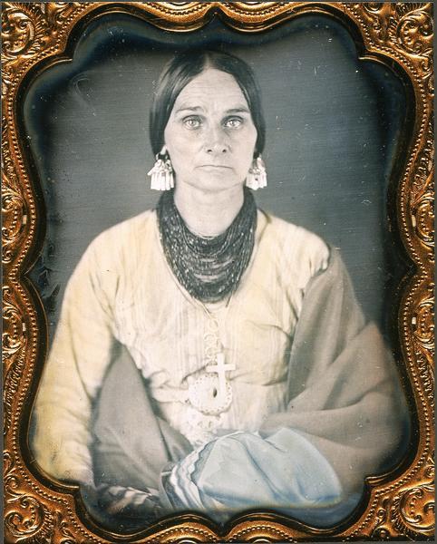 Quarter plate daguerreotype, waist-up portrait of Rachel Lawe Grignon, daughter of John Lawe and wife of Pierre Bernard Grignon. Wearing heavy bead necklace, earrings, and cross pendant, and wrapped in several blankets. Clothing and accessories are partly Native American style. Hand-tinted dress and blankets. 