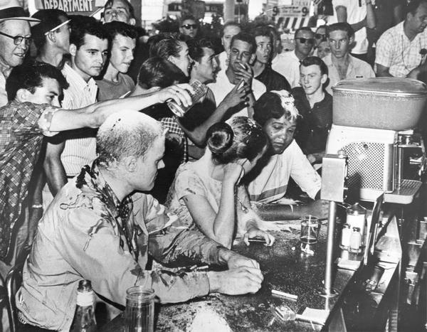 Civil rights sit-in at Woolworth's lunch counter. Seated at the counter from left to right are by John Salter, Joan Trumpauer, and Anne Moody. People pour sugar, ketchup and mustard on them in protest. Looking on as part of the crowd are Red Hydrick, seen in the upper left wearing a hat and eyeglasses, and teenager D. C. Sullivan, who is in the middle of the crowd smoking a cigarette.