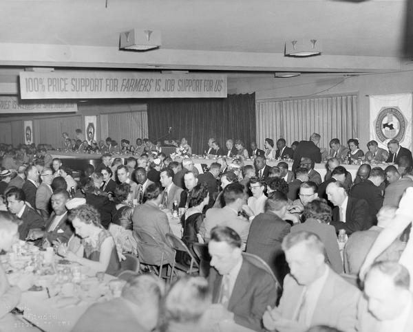 An integrated banquet hall during the Ninth Convention in Sioux City, Iowa. There is a banner overhead that reads "100% Price Support For Farmers Is Job Support For Us".