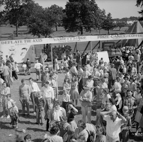 Slightly elevated view of crowd gathered at a Civilian Defense Rally. Booths are in the background, including a game booth where you can "Deflate The Axis," featuring caricatures of WWII villains to be used as target practice which is being run by the Cub Scouts, a "Variety Store" run by the Catholic Women's Club, and a "Prize Cakes & Coffee" booth run by the Women's Club.