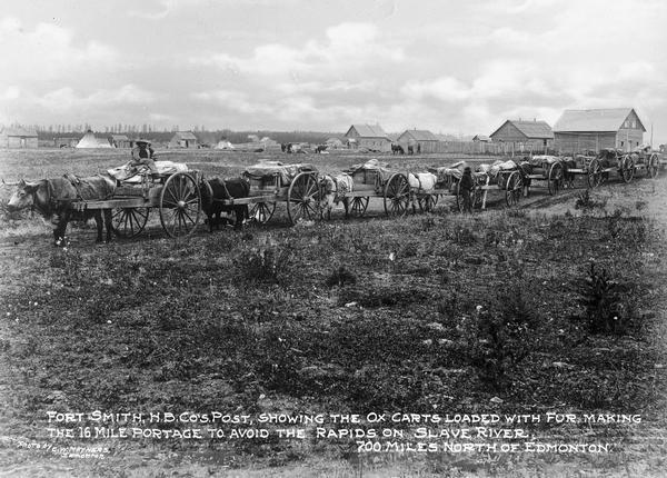 A view showing a line of ox carts loaded with furs leaving the Hudson Bay Company's Post to make the 16 mile trip to avoid the rapids on Slave River. Housing is in the background. Caption reads: "Fort Smith, H.B. Co's. Post, showing the ox carts loaded with fur, making the 16 mile portage to avoid the rapids on Slave River. 700 miles North of Edmonton." and "Photo by C.W. Mathers, Edmonton."