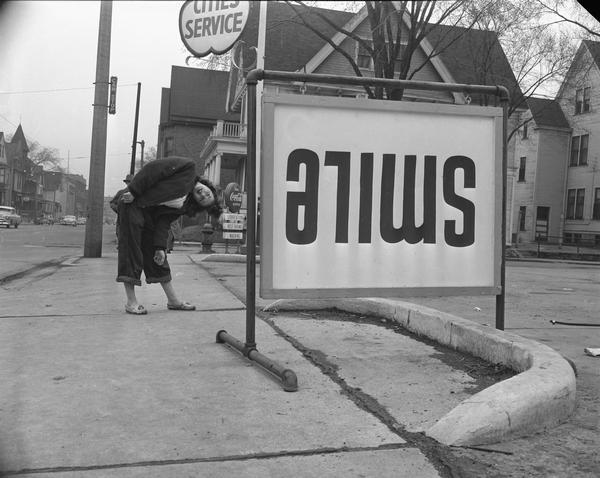 Filling station sign reading: "smile," purposely put upside down. A young woman is bending over to read it.