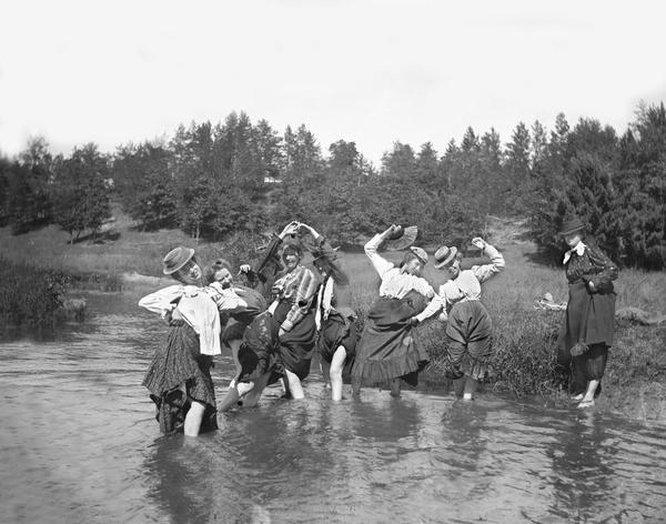 Group of women in comical poses pulling up their skirts while standing  in a stream. One woman stands on the river bank on the right. Behind them is a hill with trees.