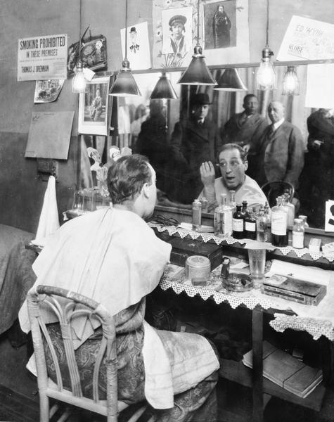 Ed Wynn is shown in his dressing room applying makeup at the mirror. Reflected in the mirror left to right are Wynn's manager Fred R. Zweifal, Wynn's valet, and George W. Lederer.