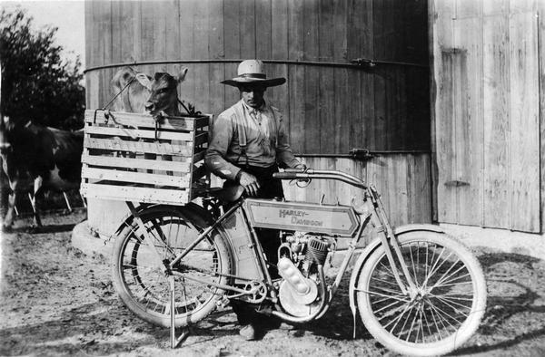 A man poses next to his 1912 model Harley-Davidson motorcycle with a young calf in a crate strapped on the back of the motorcycle. Behind the man is a silo and barn.