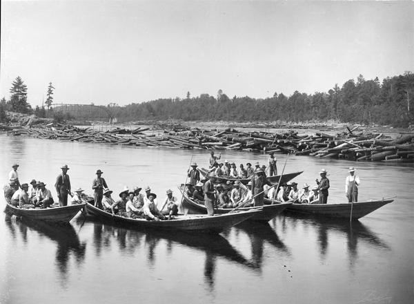 Log driving crew on river in five bateaux. In the background on the left is a bridge.