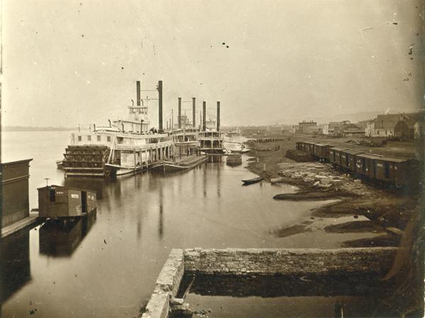 The sternwheel packet, <i>Arkansas</i>, next to a barge with other steamboats at the levee in Winona between 1870 and 1880. Railway cars are on a siding in the yard next to the levee.