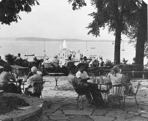 Summertime view of people at tables on University of Wisconsin-Madison Memorial Union terrace, looking out to canoes and sailboats on the lake. In the background people are wearing bathing suits and standing on a pier.