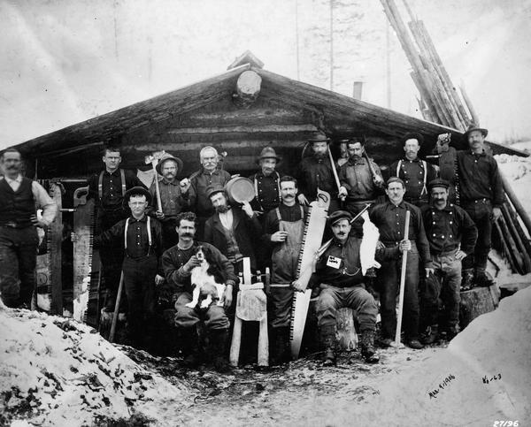 Camp Montana loggers pose in front of camp building with their tools.  Includes a dog sitting on the lap of one logger.