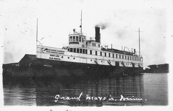 The screw passenger and freight vessel, <i>America</i>, docked. Sign on boat reads: "United States and Dominion Transportation Company, Booth Line, U.S. Mail, America." Caption reads: "Grand Mareis, Minn."