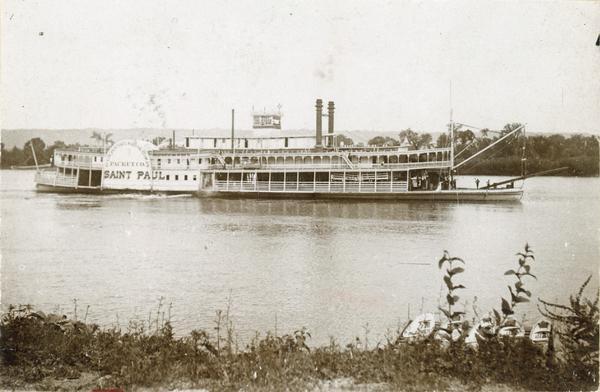 The sidewheel excursion, <i>Saint Paul</i>, on the Mississippi River near Lansing, Iowa. Later named the <i>Senator</i>. Small boats are line up on a pier near the bank in the foreground. 