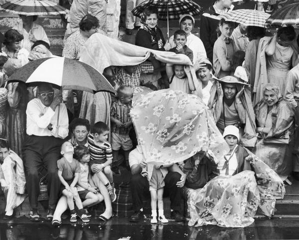 A group of people watching a parade along a sidewalk in the rain.