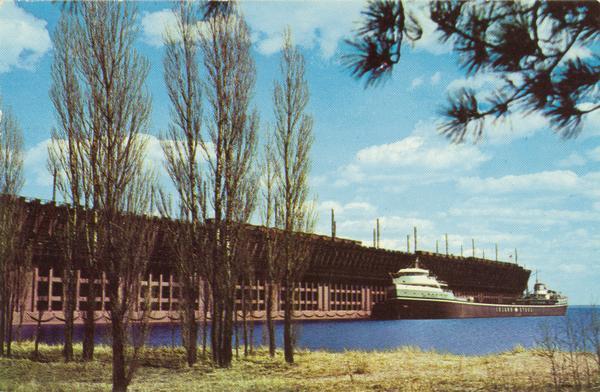 The screw bulk freighter, Wilfred Sykes at L.S. and I. Ore Dock in Marquette, Michigan about 1960. Lake shore and trees are in the foreground.