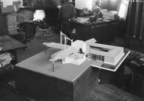 Architectural model of a village-type gasoline station designed by Frank Lloyd Wright on display at Taliesin. Taliesin is located in the vicinity of Spring Green, Wisconsin. Men are in the background near a fire in the fireplace.