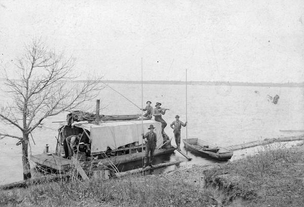 Five lumberjacks, one of whom is playing the fiddle, pose by a docked riverboat with a rowboat nearby.